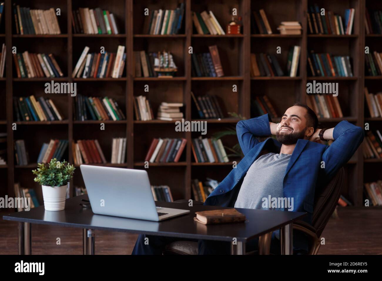 Giovane uomo d'affari con portacolori di successo in una tuta blu nella biblioteca dell'ufficio. Scrivania dell'uomo d'affari con laptop. Tempo di rilassarsi Foto Stock