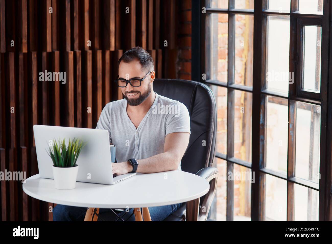 Un uomo d'affari freelance in una t-shirt bianca e gli occhiali sta lavorando su un laptop. Digitando su una tastiera del computer in un accogliente ufficio coworking Foto Stock