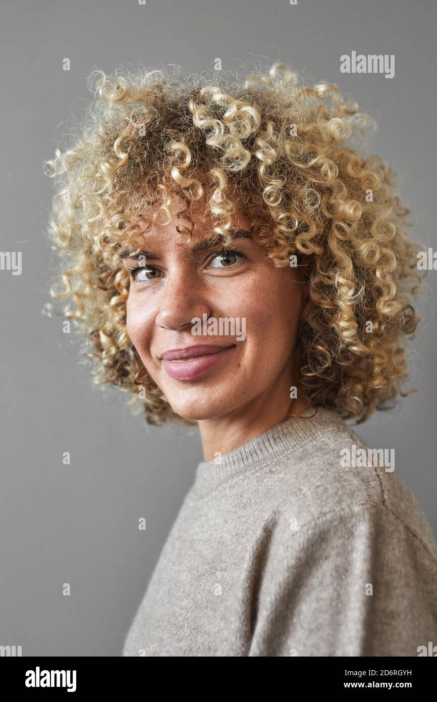 Ritratto verticale di una donna sorridente dai capelli ricci che guarda la fotocamera mentre si posa su sfondo grigio, concetto di bellezza unico Foto Stock