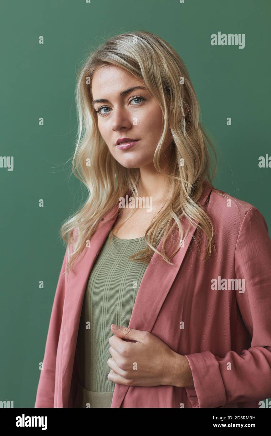 Verticale vita in su ritratto di elegante donna bionda che indossa polveroso giacca rosa e guardando la fotocamera mentre si posa contro il verde sfondo in studio Foto Stock