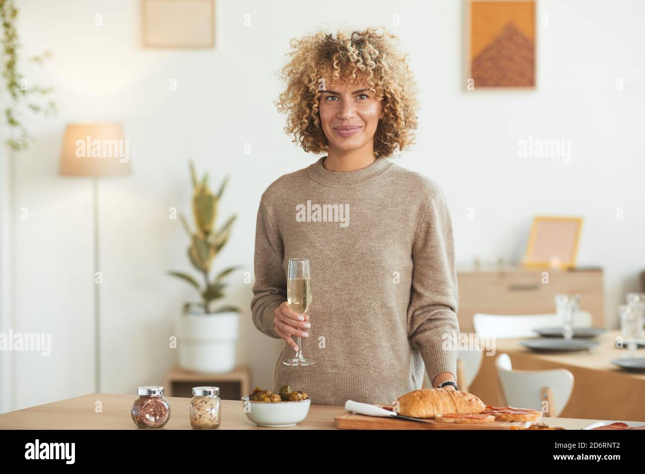 Ritratto in vita di una donna mista contemporanea che tiene un bicchiere di champagne e guarda la macchina fotografica mentre si cucina per la cena in casa, spazio copia Foto Stock