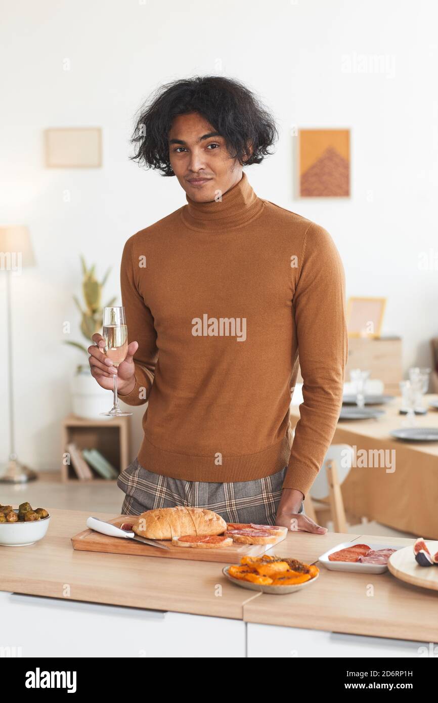 Ritratto verticale in vita di un uomo contemporaneo di razza mista con champagne vetro e guardando la macchina fotografica mentre cucinano per la festa di cena all'interno Foto Stock