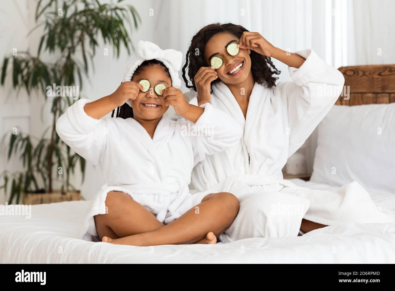 Giocosa madre nera e figlia in accappatoi che applicano i pezzi del cetriolo agli occhi Foto Stock