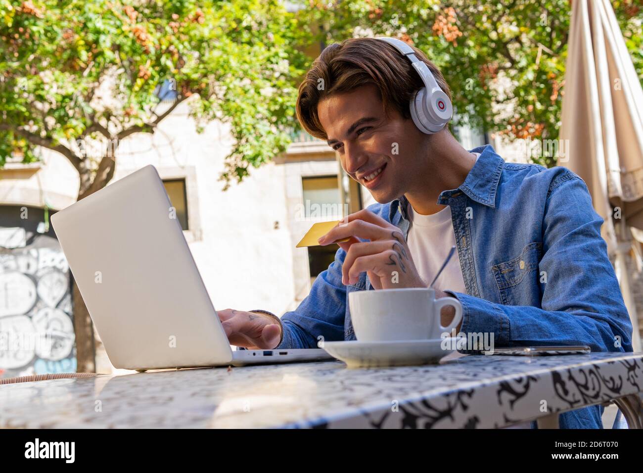 Giovane compratore di contenuti maschile in cuffie con carta di debito guardare netbook mentre si acquista online in strada Foto Stock