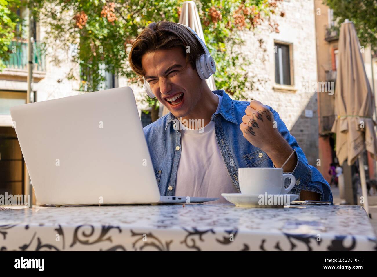 Il giovane maschio è stato molto felice di avere le cuffie wireless e di avere notizie incredibili laptop e festeggiando la vittoria mentre si siede a tavola con la tazza di caffè sul caffè Foto Stock