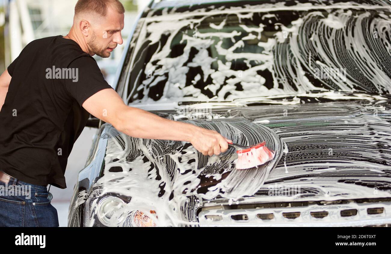 Un giovane serio che indossa una t-shirt nera sta lavando la sua auto con una spazzola e sapone all'aperto, vista laterale, primo piano Foto Stock