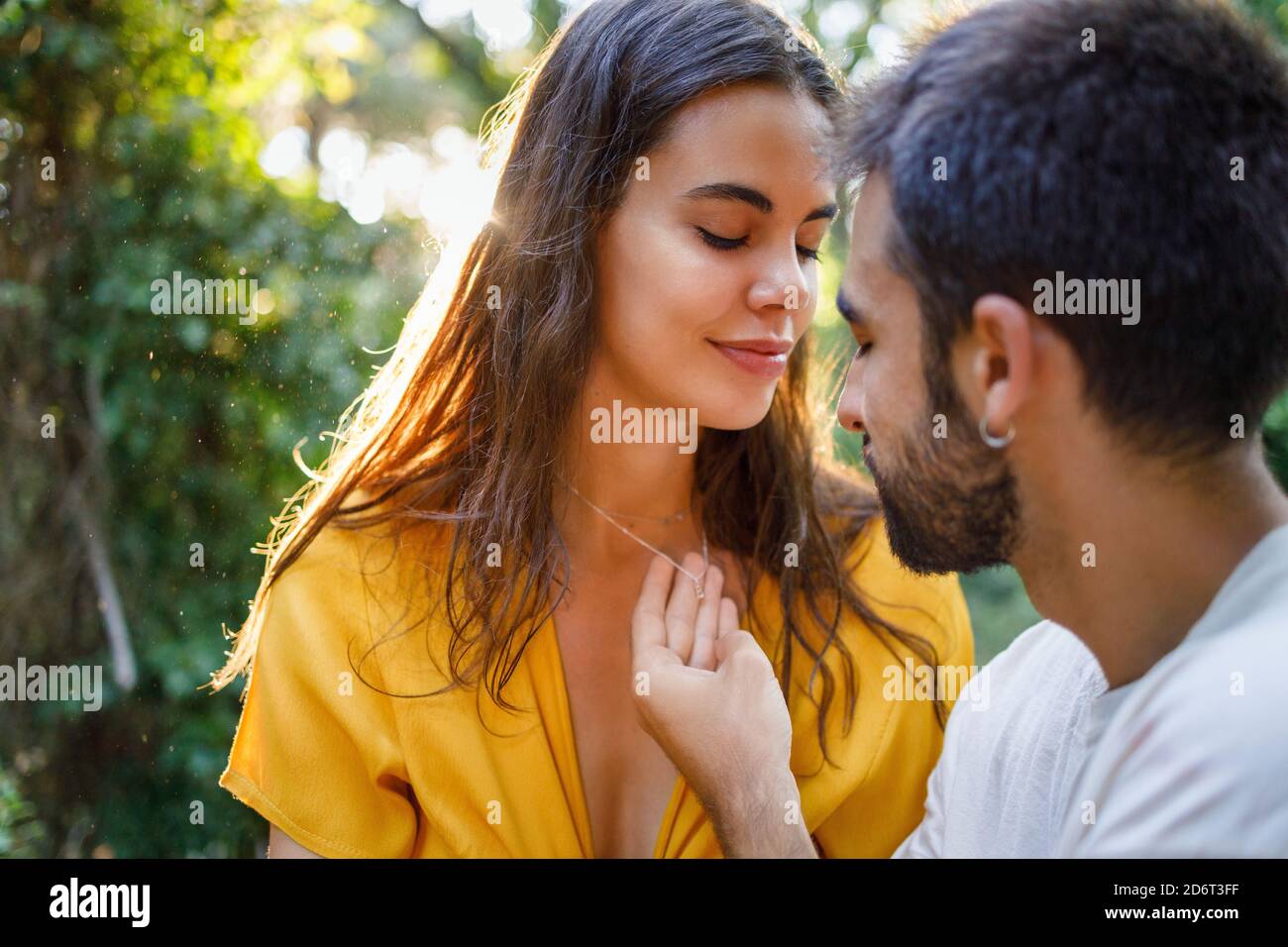 Vista laterale di giovane ragazzo etnico bearded che tocca la collana di ragazza felice mentre si rilassa insieme in giardino verde sul sole giorno Foto Stock