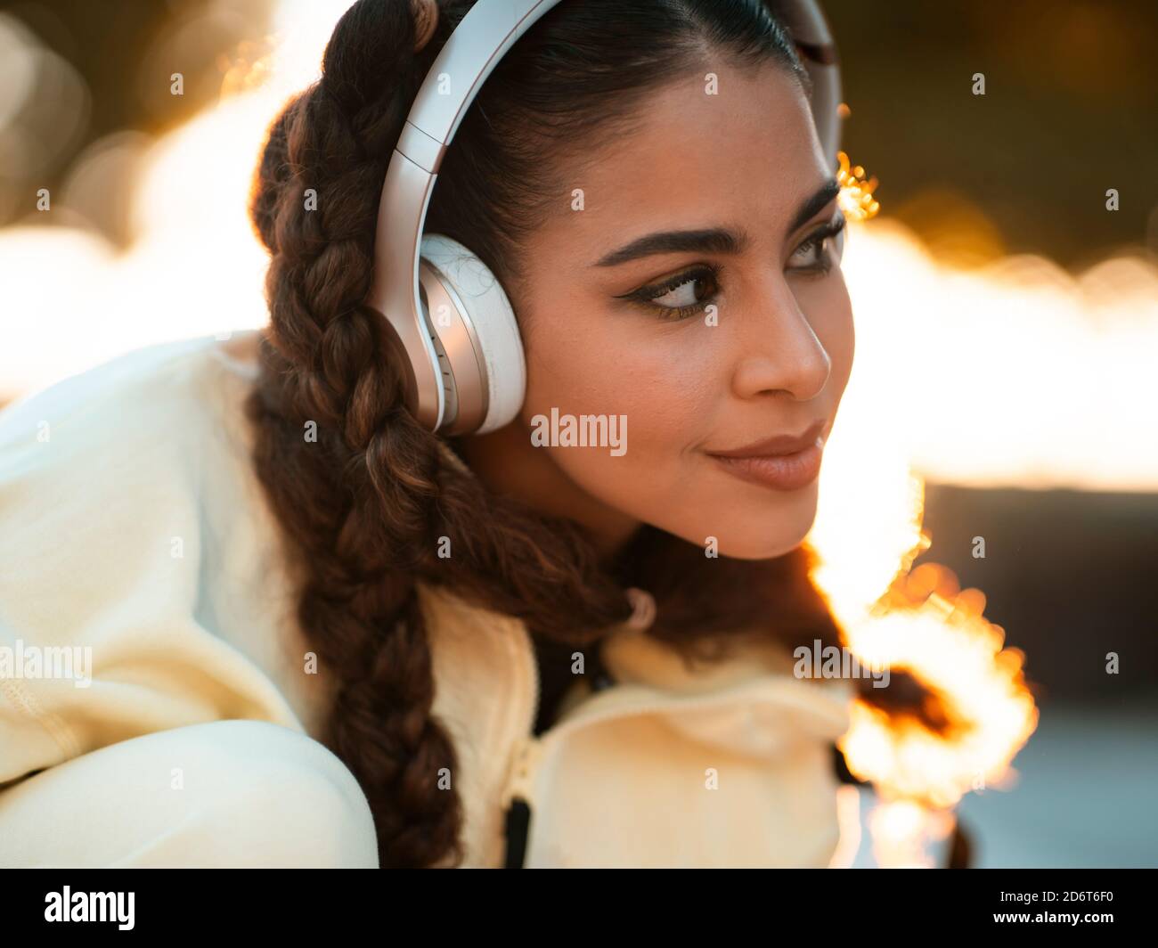 Giovane donna etnica cropped positivo in elegante sportswear che cavalcano la longboard e ascoltare la musica in cuffia al tramonto Foto Stock