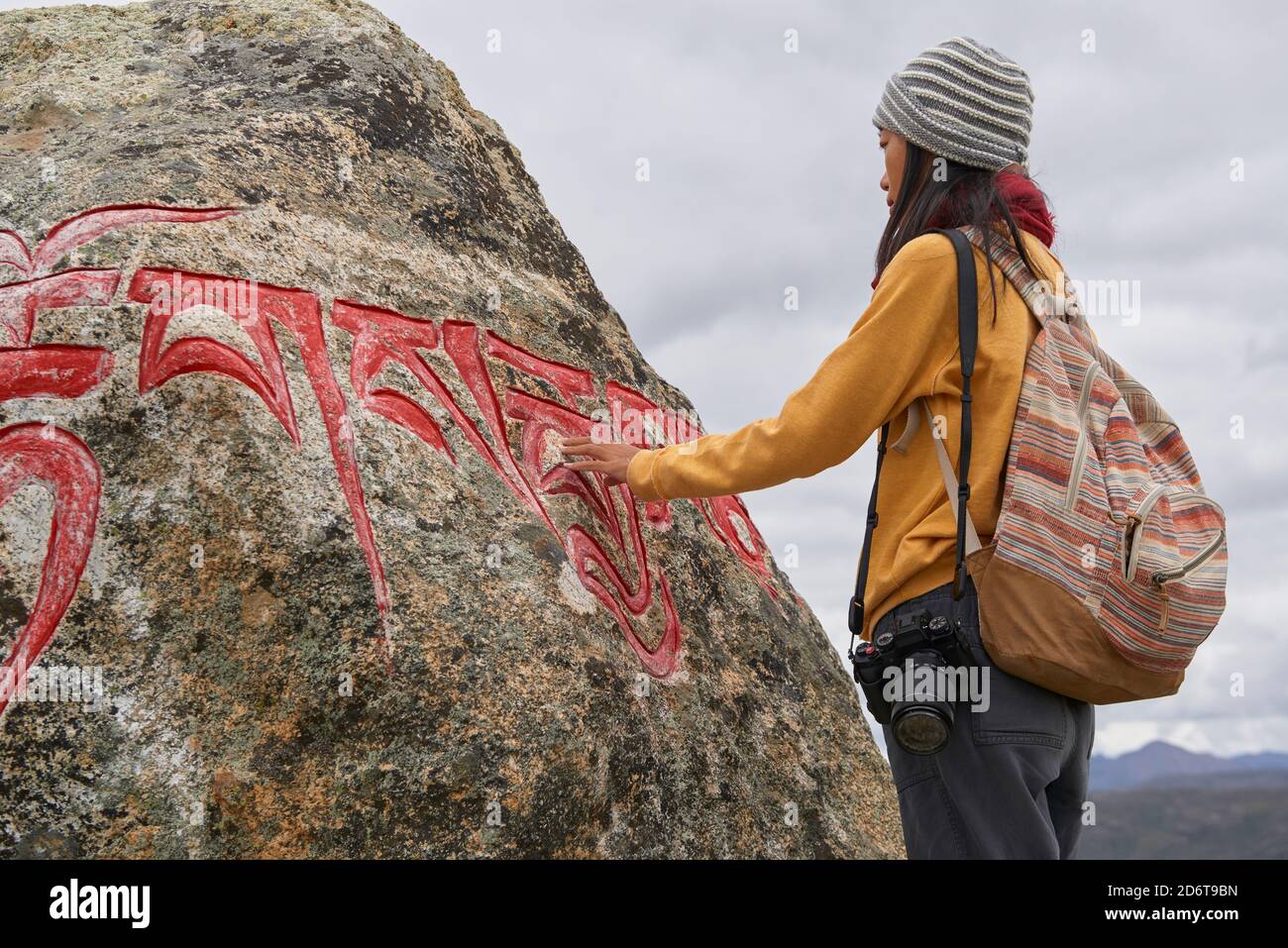 Vista laterale del corpo completo del turista femminile in abiti caldi e cappello guardando grande pietra con Om mani Padme Hum mantra sulla superficie nel villaggio di Baiyu in Foto Stock