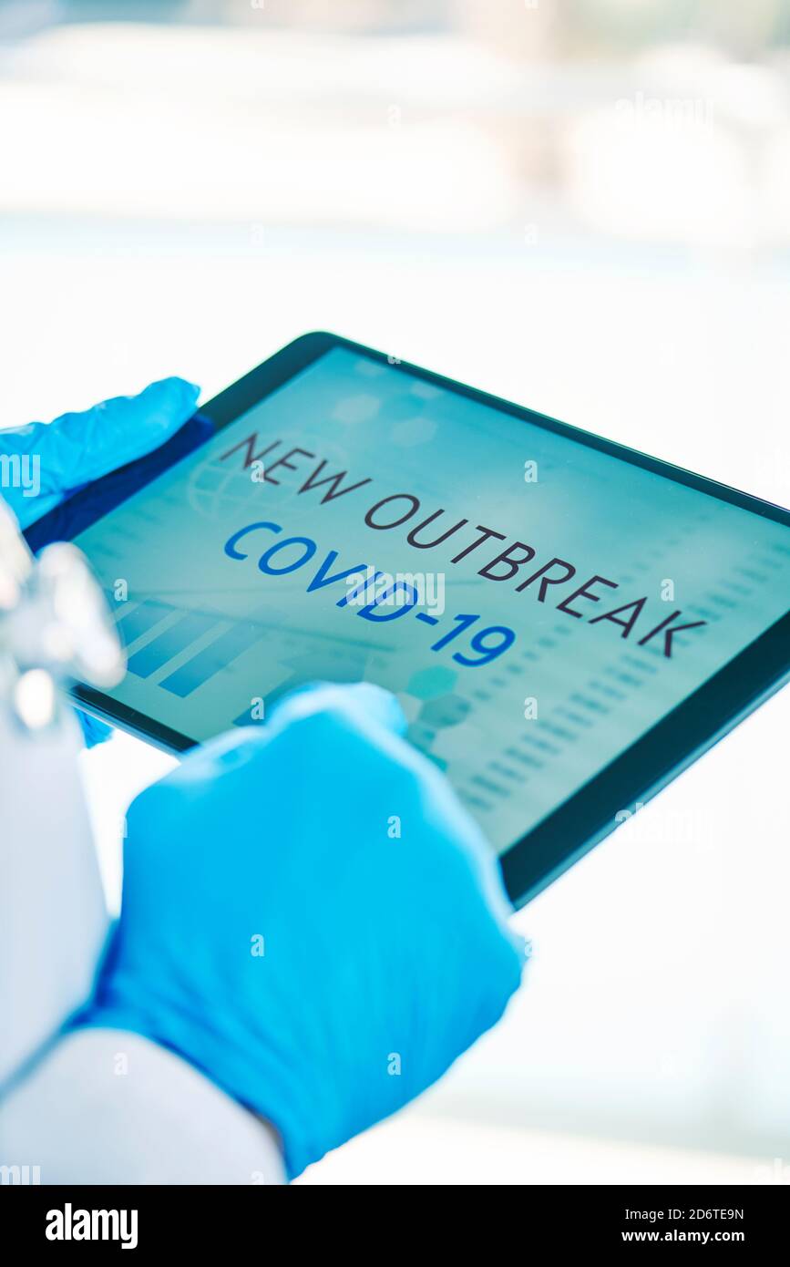 primo piano di un medico, indossando guanti chirurgici blu, avendo una tavoletta digitale in mano con il testo covid-19 nuovo focolaio nel suo schermo Foto Stock
