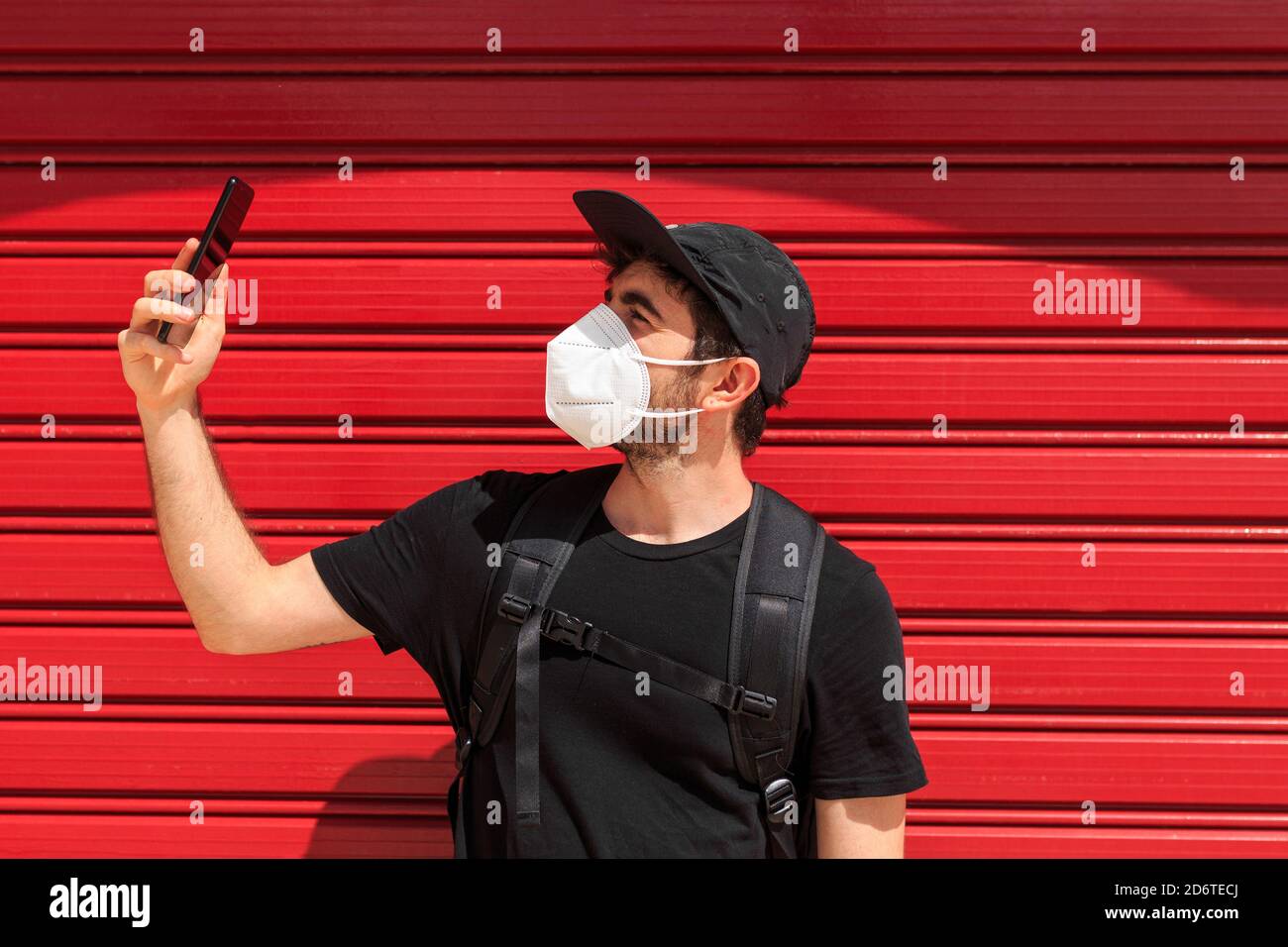Giovane maschio in maschera respiratoria prendendo selfie sul cellulare vicino Muro rosso in piedi sulla strada durante la pandemia COVID 19 Foto Stock