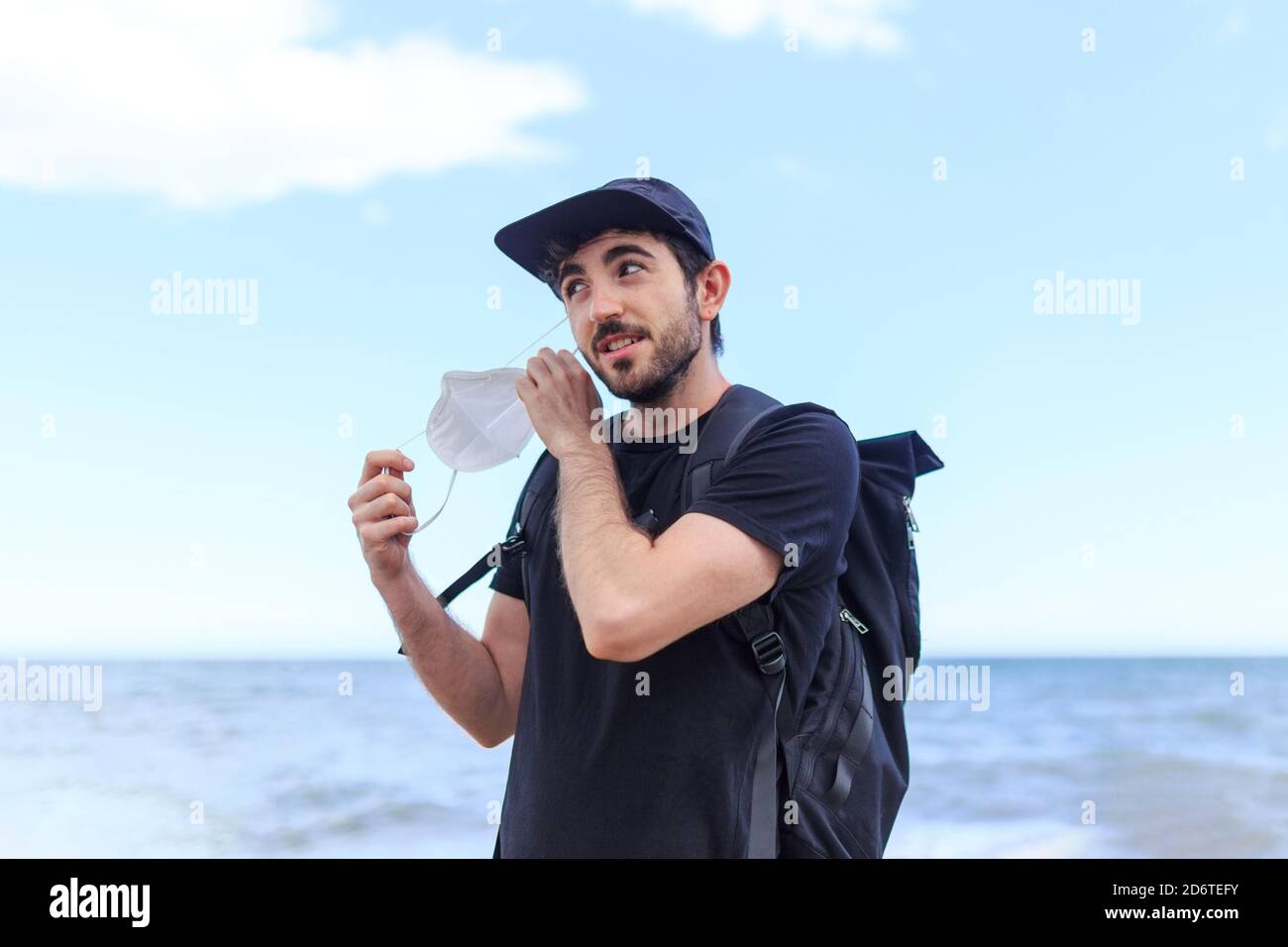 Sorridente portatore maschio in cappello con zaino in piedi prendendo off maschera protettiva in piedi dietro l'oceano sotto il cielo blu nuvoloso Foto Stock