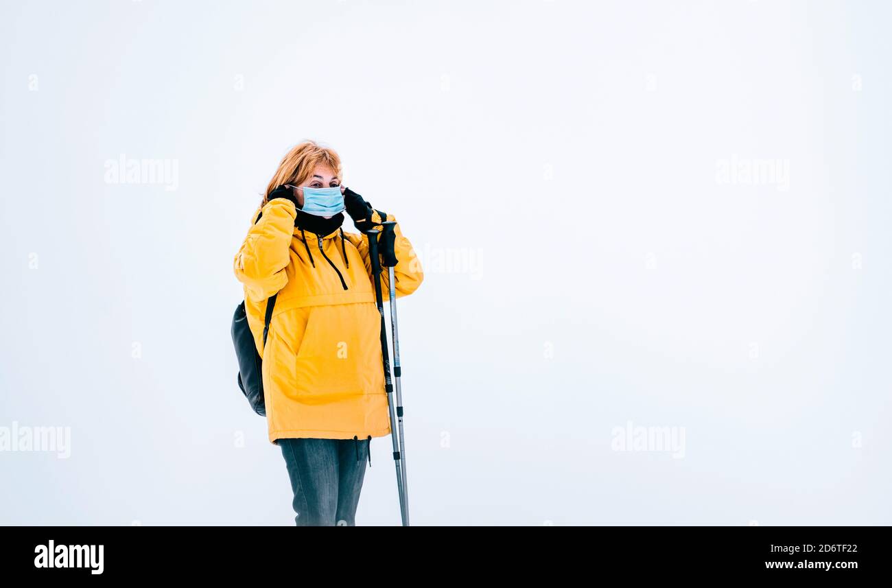 Donna escursionista con outerwear giallo e maschera protettiva che guarda via mentre si è in piedi con bastoni da trekking in montagne innevate Foto Stock