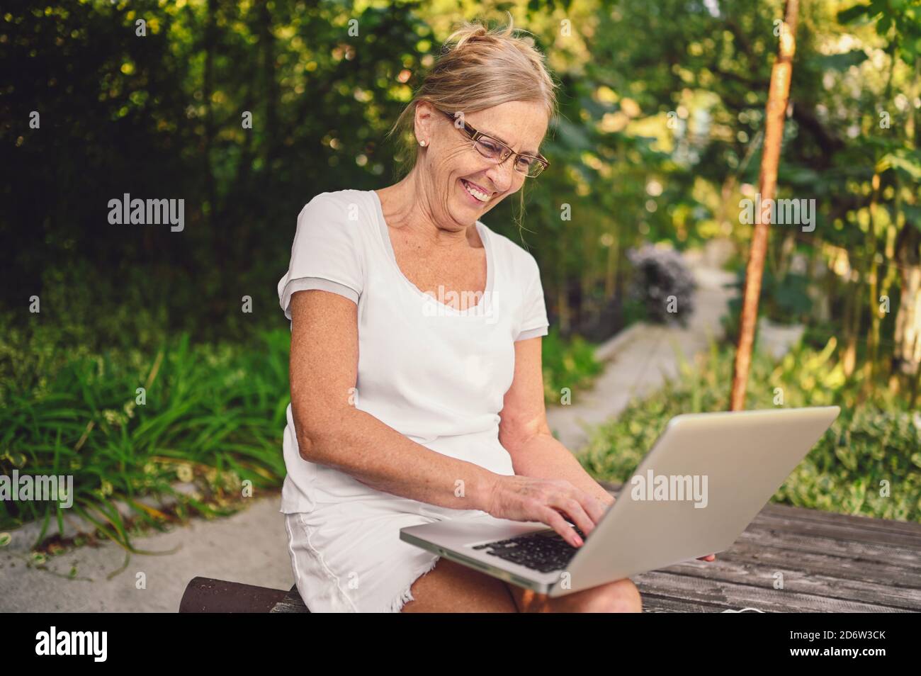 Tecnologia, concetto di gente anziana - anziana anziana anziana felice che lavora in linea con il computer portatile all'aperto nel giardino. Lavoro a distanza, formazione a distanza. Foto Stock