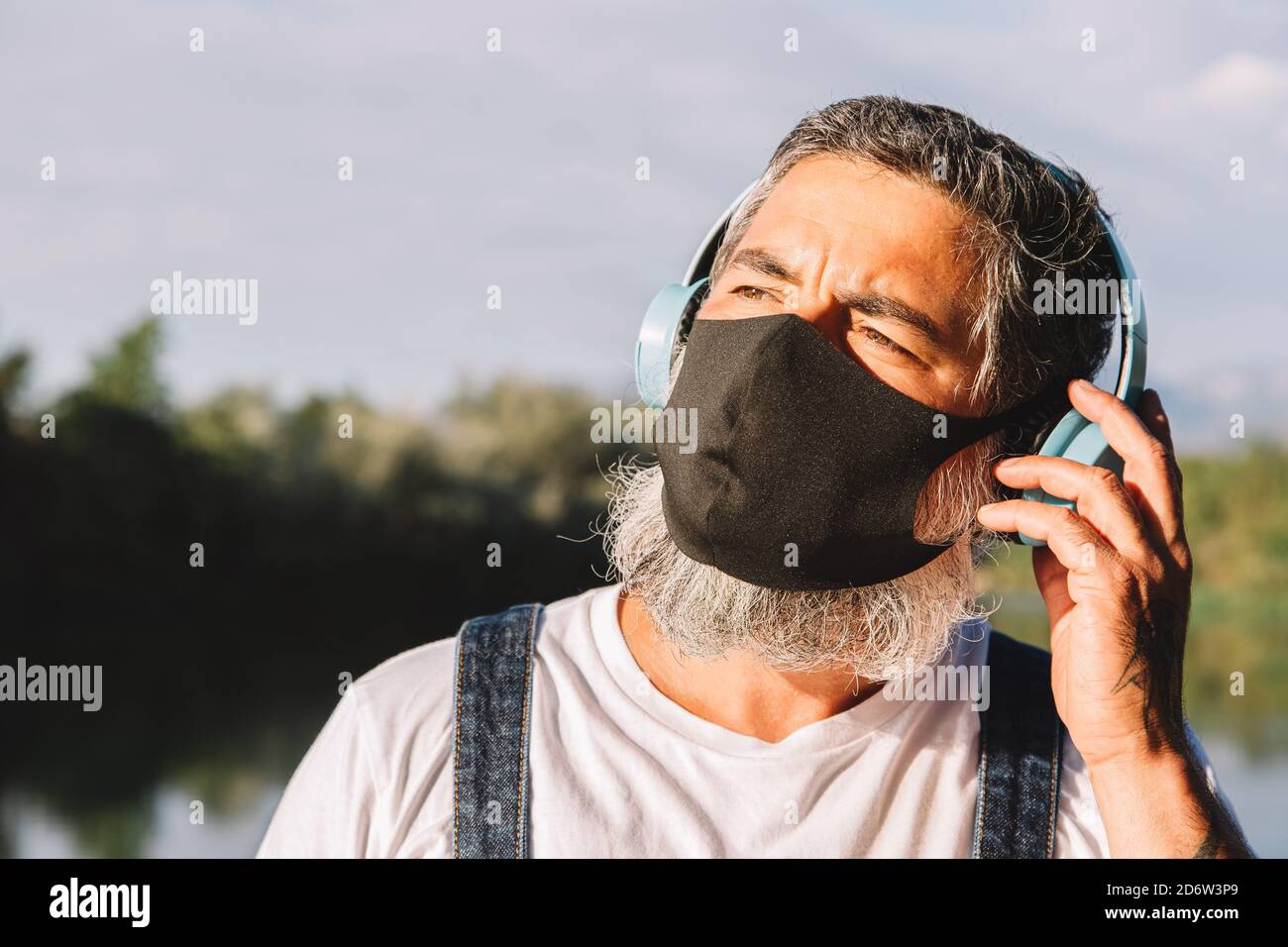 Uomo maturo con bearded che indossa cuffie e maschera per il viso grazie al coronavirus all'aperto godendo della luce del sole. Foto Stock