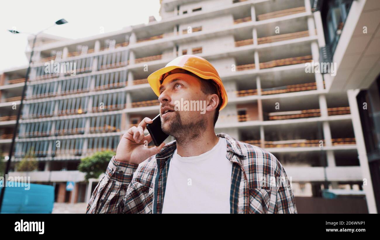 Uomo caucasico lavoratrice di costruzione in cappello arancione e camicia plaid che parla al telefono al cantiere. Tema dell'architettura. Professione maschile Foto Stock