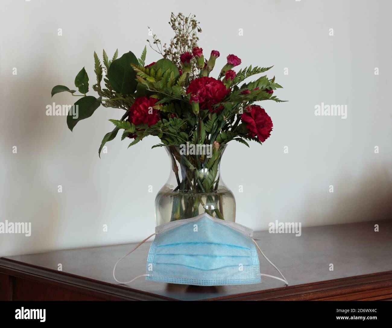 una maschera blu posta di fronte a un vaso di fiori di garofano rosso su un tavolo di legno scuro accanto su una parete bianca con spazio per la copia Foto Stock