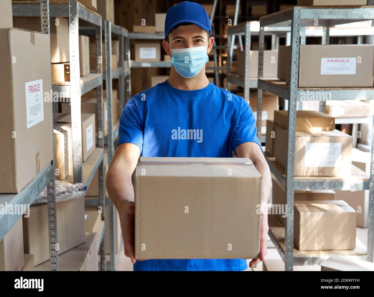 Corriere maschio indossando maschera facciale tenendo scatola pacchi in magazzino, ritratto. Foto Stock