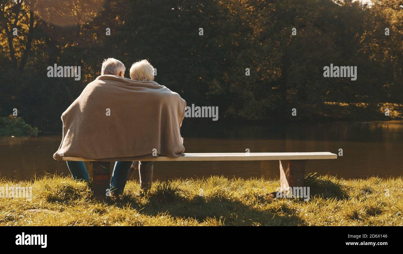Anziana coppia di capelli grigi che si siede sulla panchina vicino al fiume che copre con coperta il giorno d'autunno. Romanticismo e vecchiaia. Foto di alta qualità Foto Stock