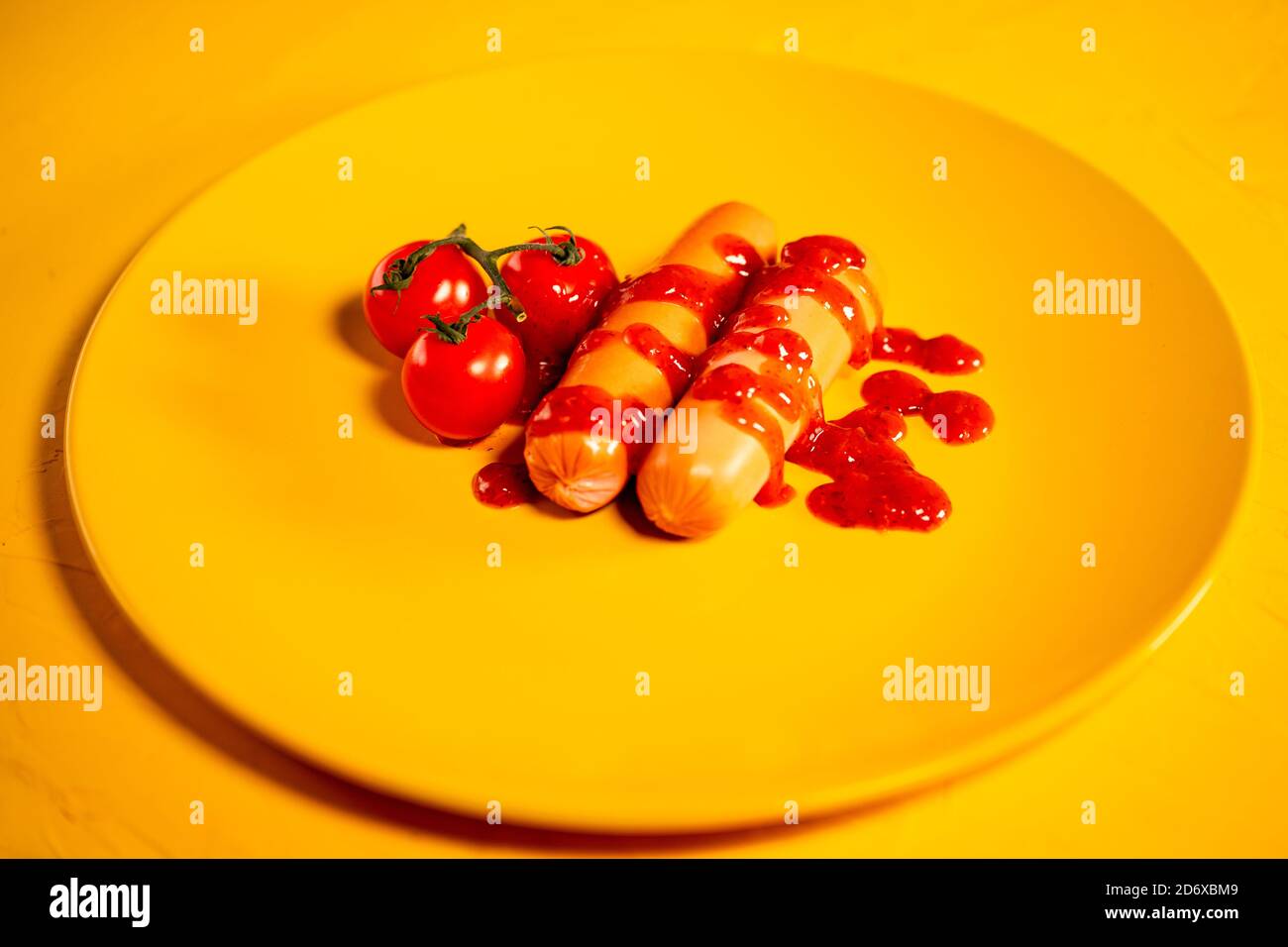 Primo piano di salsicce in salsa con pomodori ciliegini su piatto giallo. Appetitosi frankfurters con piccoli pomodori rossi e ketchup su sfondo giallo. Foto Stock