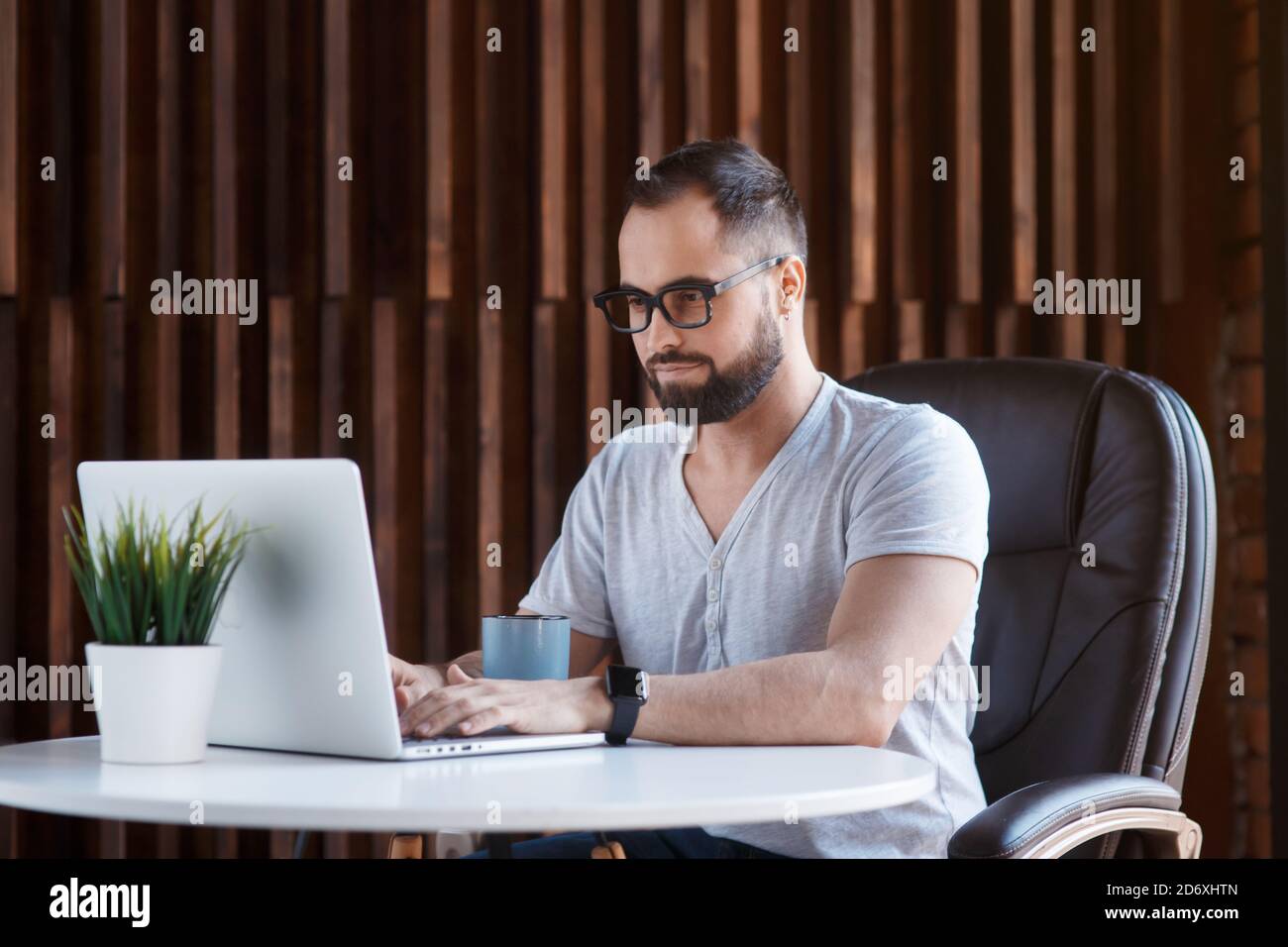 Un uomo d'affari freelance in una t-shirt bianca e gli occhiali sta lavorando su un laptop. Digitando su una tastiera del computer in un accogliente ufficio coworking. Foto Stock