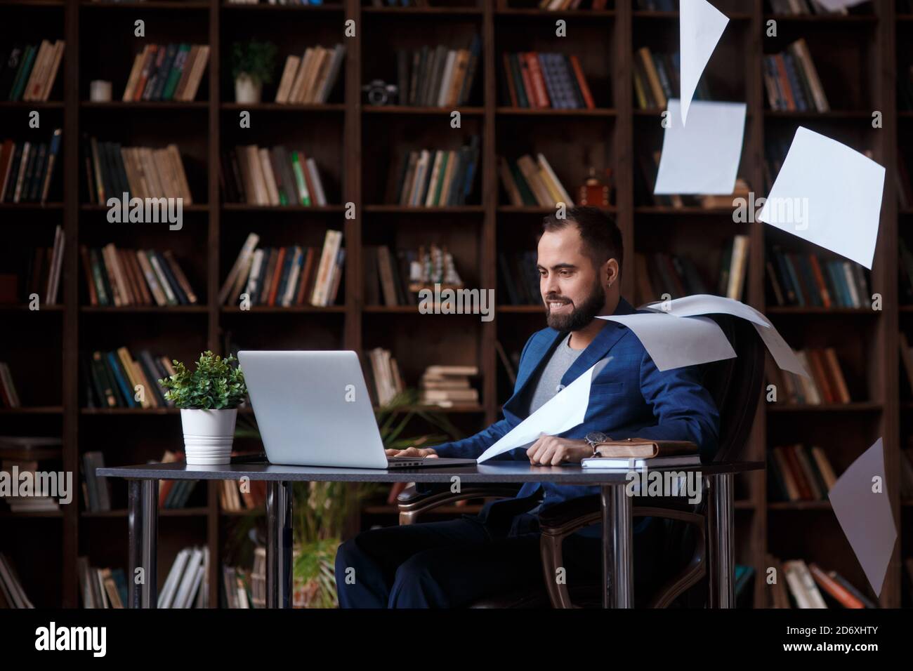 arrabbiato pazzo businessman bearded in biblioteca ufficio seduto sul tavolo di fronte laptop. l'uomo getta i documenti in rabbia. Uomo d'affari in male Foto Stock