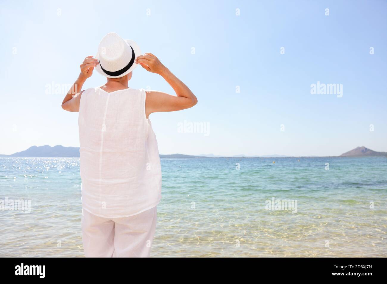 Donna felice anziana si ritirò godendo il sole che indossa il cappello sulla vacanza di spiaggia dei Caraibi. Viaggio turistico in piedi al mare e cielo limpido. Vista posteriore dell'innesto femmina Foto Stock