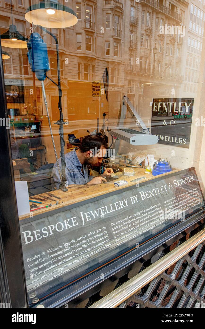 Finestra di Bentley e Skinner, gioiellerie di alta classe a Piccadilly, Londra; un uomo sta riparando gioielli utilizzando una lente di ingrandimento Foto Stock