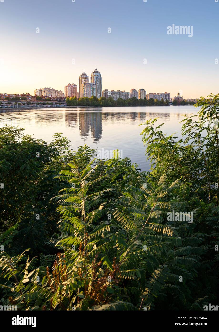 Edifici alti Obolon vicino al fiume Dnieper a Kiev, Ucraina. Cielo blu chiaro e riflessione in acqua. Varie piante e vegetazione nello straniero Foto Stock