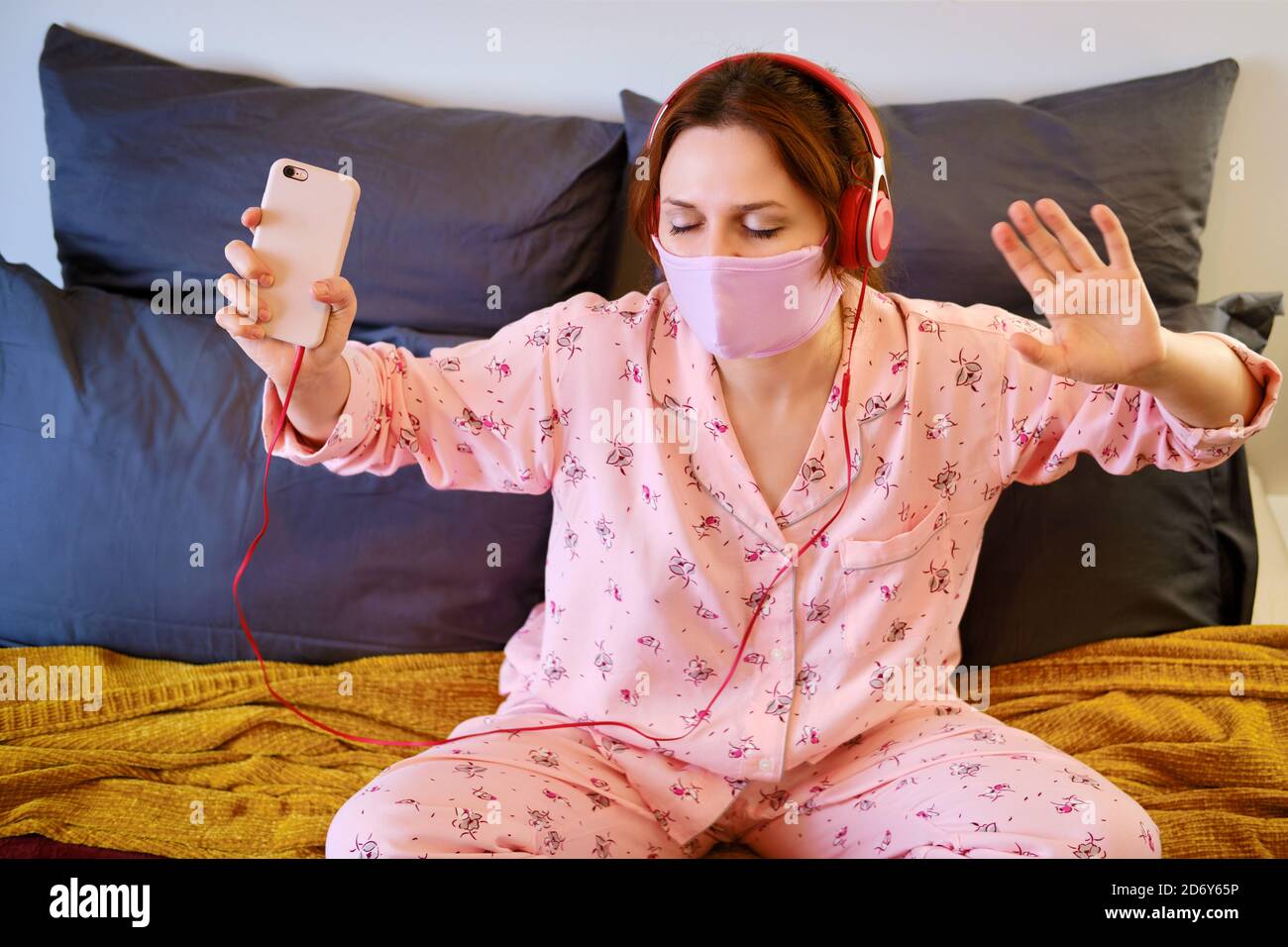 Una donna ascolta musica e balli mentre si siede sul letto durante una malattia con il virus dell'influenza covid-19. Ragazza in pigiama, cuffie e un protettori Foto Stock