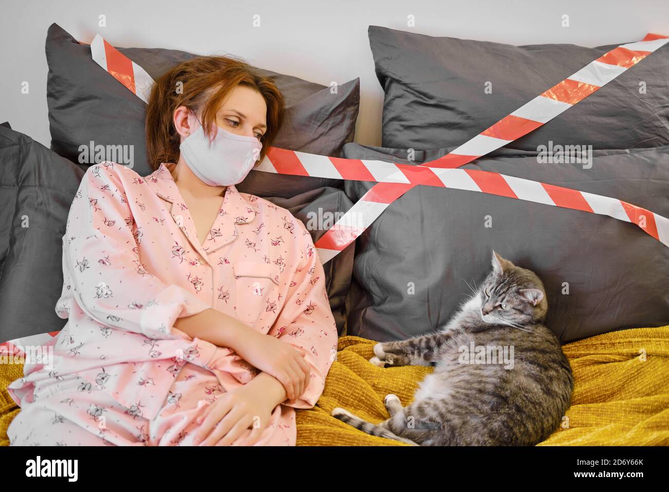 Una donna in una maschera medica casalinga si trova con un gatto durante una malattia del coronavirus, concetto. Ragazza dai capelli rossi in pigiama su un letto con un nastro di arresto durante Foto Stock