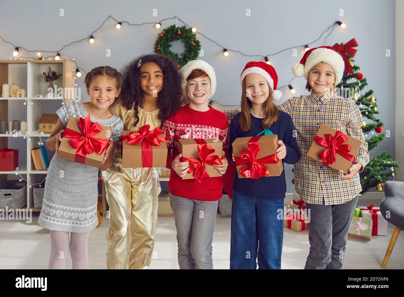 Felici i bambini diversi che tengono i regali di Natale e guardando la macchina fotografica in piedi in un accogliente soggiorno Foto Stock