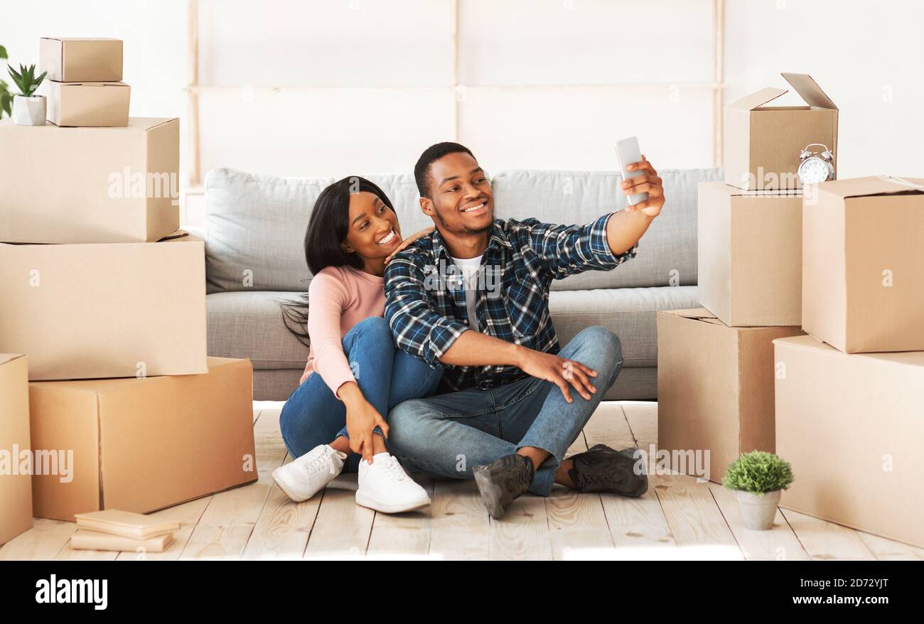 Sorridente ragazzo nero e la sua ragazza che si spostano a casa loro insieme, prendendo selfie tra scatole di cartone, panorama Foto Stock