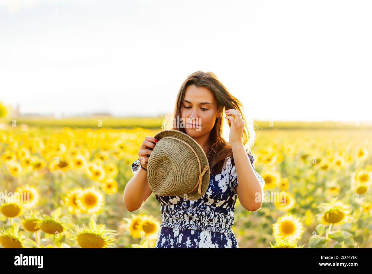 Giovane bella donna bruna in posa nel suo vestito designer in un campo di girasoli e indossare un cappello Foto Stock