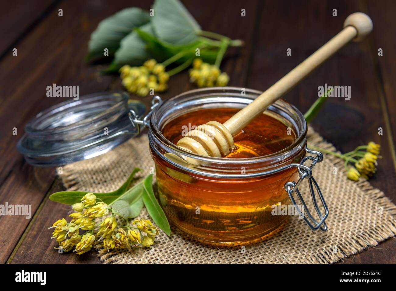 Vaso con miele di tiglio con bastone per miele e fiori di tiglio freschi su un tavolo di legno. Foto Stock