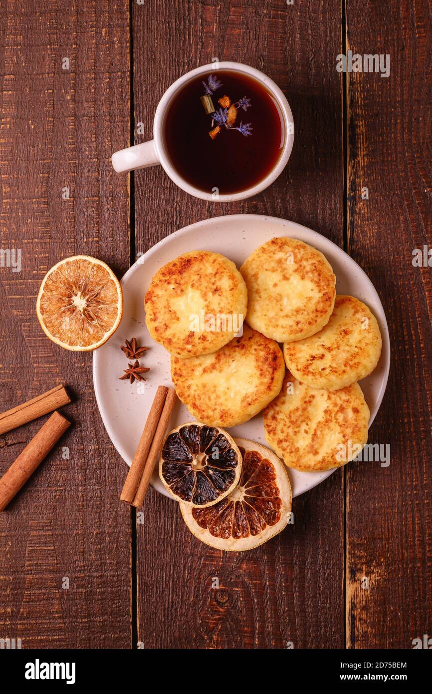 Frittelle di formaggio cottage con tè nero aromatico caldo, umore di prima colazione di Natale con anice, cannella e agrumi secchi su sfondo di legno, vista dall'alto Foto Stock