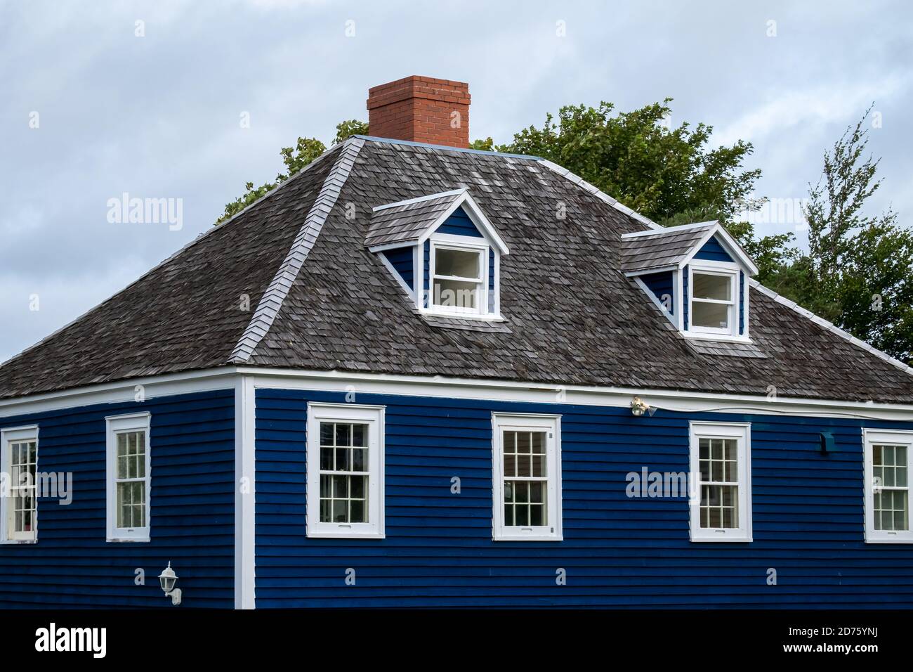 Tetto vintage hip con due dormitori, tetto in cedro, camino in mattoni rossi, pannello di copertura blu, finestre doppie appese, e rifiniture bianche sull'edificio. Foto Stock