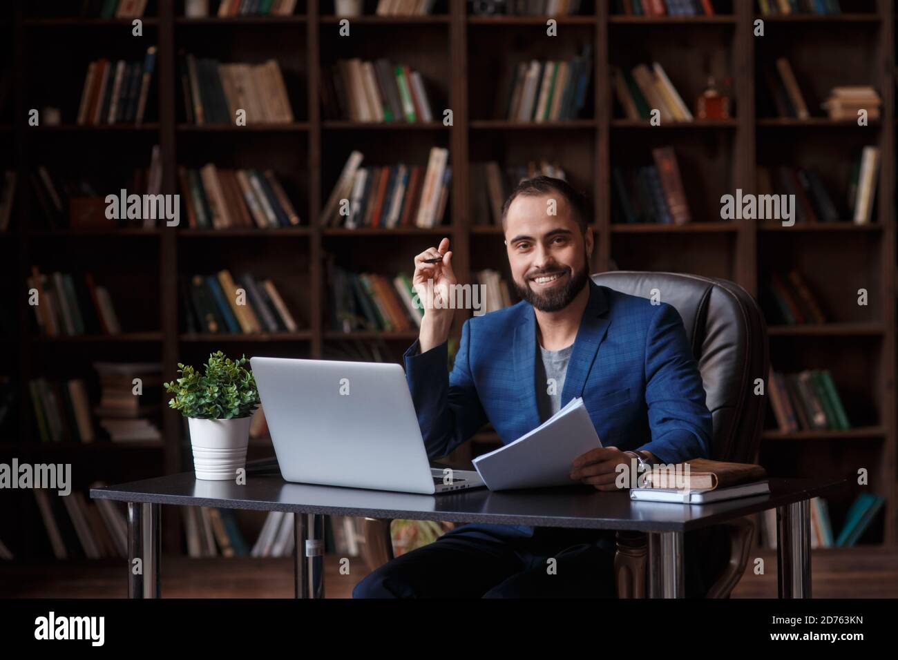 Un giovane uomo d'affari sorride e guarda al telaio tra i libri nella biblioteca a un tavolo in parte anteriore di un computer portatile Foto Stock