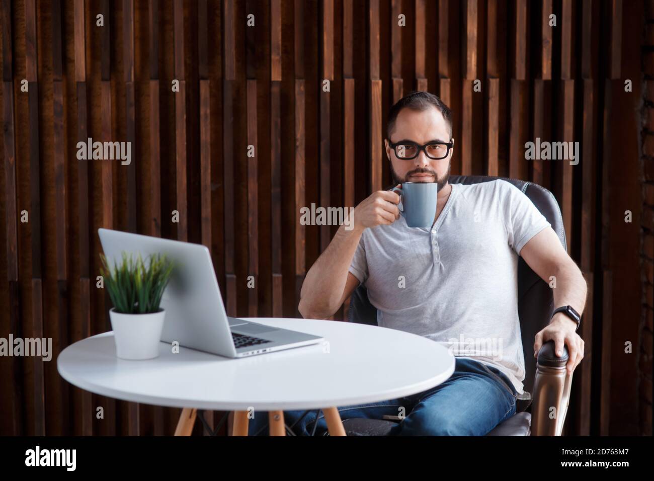 Giovane uomo d'affari sopportato di successo freelancer in ufficio coworking. Programmatore uomo felice sviluppatore in una t shirt bianca sorridente e bere caffè a. Foto Stock