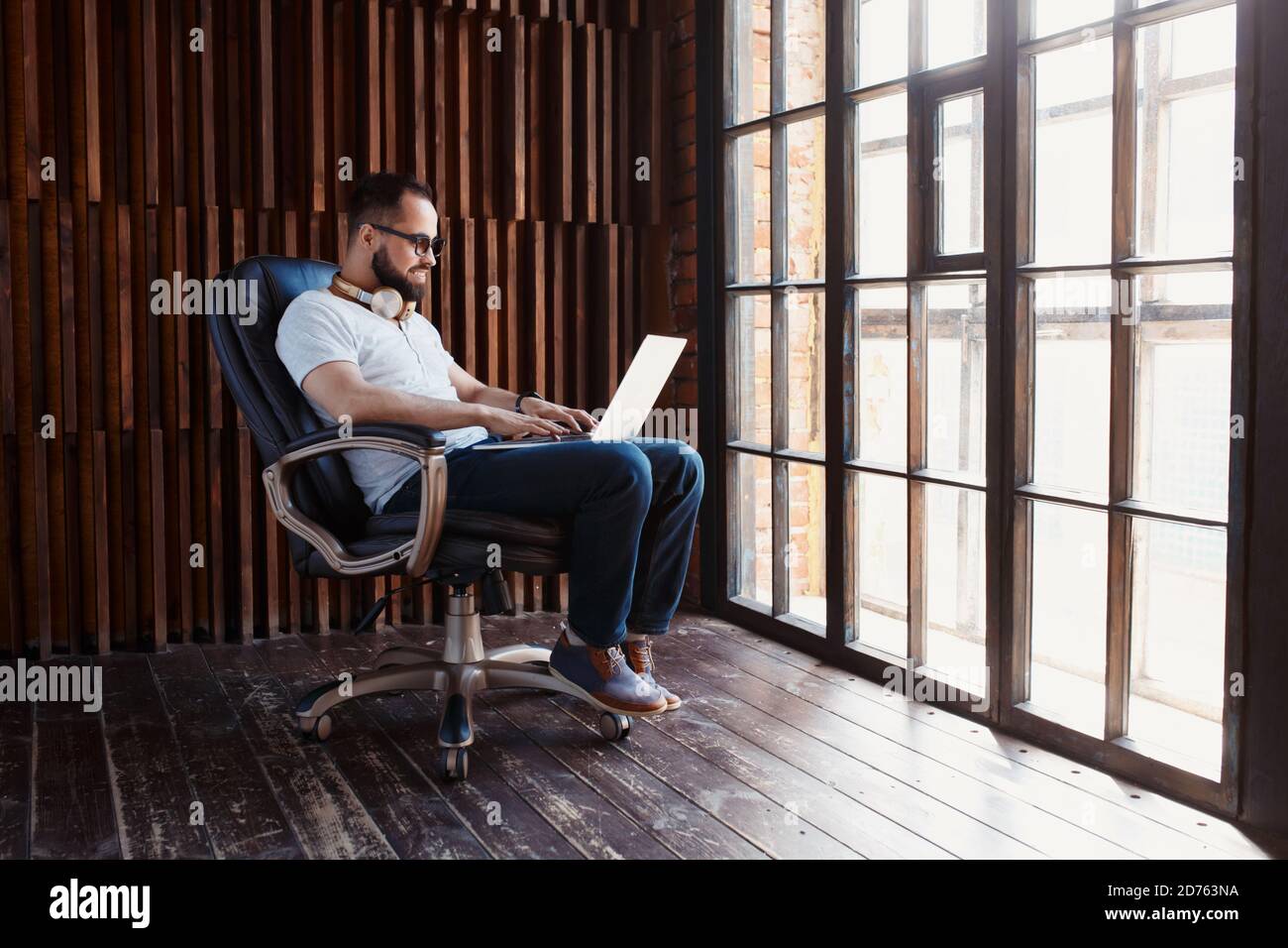 Un uomo d'affari freelance in una t-shirt bianca e gli occhiali sta lavorando su un laptop. Digita il testo sulla tastiera del computer seduto in una sedia in un ufficio accogliente Foto Stock