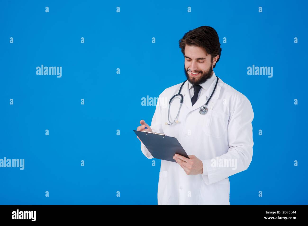 un giovane medico con la bearded in un cappotto medico bianco sopra uno sfondo blu scrive i reclami del paziente su un tablet con documenti e medici Foto Stock