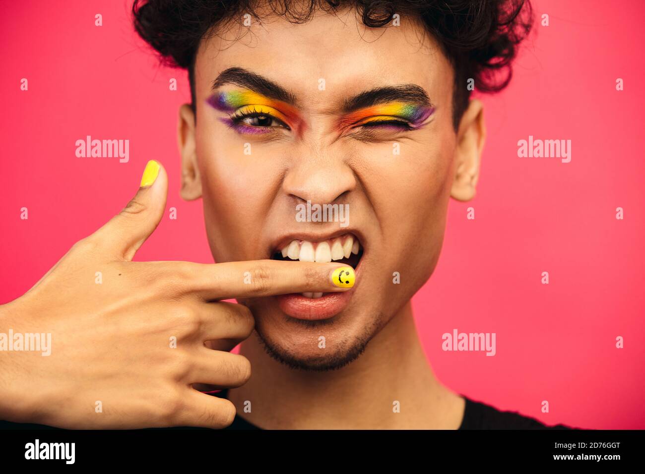 Primo piano di un uomo transgender che gli mordicca il dito e gli inzidisce un occhio. Uomo fluido di genere che indossa arcobaleno colorato occhio ombra e sorridente viso unghie. Foto Stock