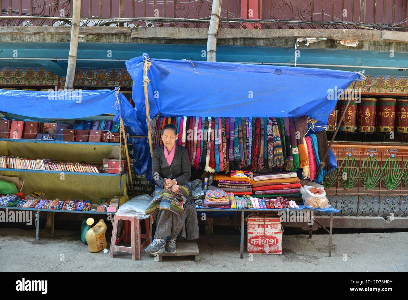 INDIA, Mcleod-Ganj. Donna venditore di souvenir nella sua piccola stalla di fronte a una fila di mulini di preghiera Foto Stock