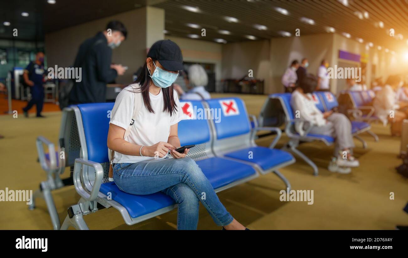 Turista asiatico con telefono cellulare, indossare maschera igienica, sedersi su sedia con divaricamento sociale per prevenire pandemia durante il viaggio al terminal dell'aeroporto. Nuovo Foto Stock