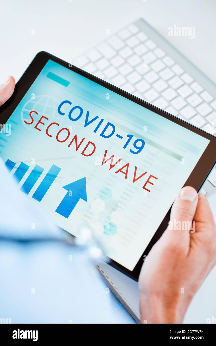 primo piano di un medico, seduto alla sua scrivania, con un tablet digitale in mano con il testo covid-19 seconda onda nel suo schermo Foto Stock