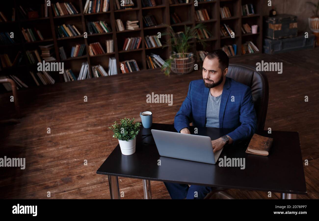 Un giovane uomo d'affari lavora su un computer portatile in un lussuoso ufficio nella biblioteca. Un uomo barbuto e allegro in un vestito blu a una scrivania sta digitando su una tastiera Foto Stock