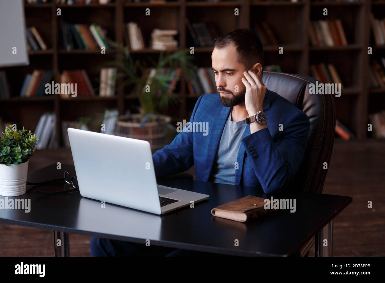 Un giovane uomo d'affari con gli occhiali sta lavorando su un computer portatile in un lussuoso ufficio nella biblioteca. Un uomo bearded in un vestito blu ad una scrivania sta digitando su un Foto Stock