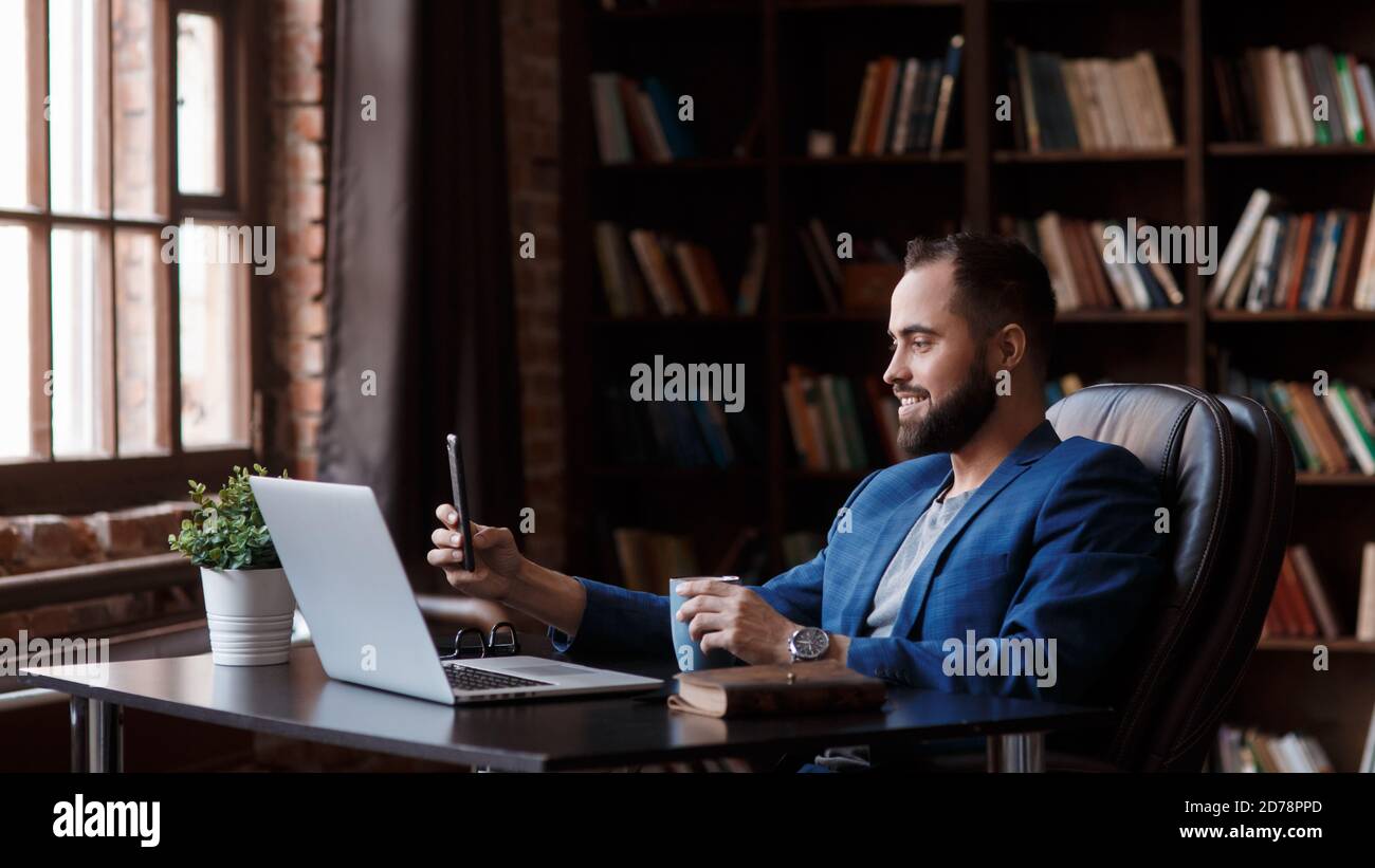 Un giovane uomo d'affari in una tuta blu nell'ufficio della biblioteca sta parlando su un collegamento video sul telefono. Lo spazio di lavoro dell'uomo d'affari - Foto Stock