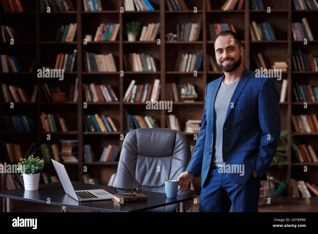 Uomo d'affari giovane in un vestito blu nella biblioteca dell'ufficio. Un uomo si trova sul desktop con un notebook Foto Stock