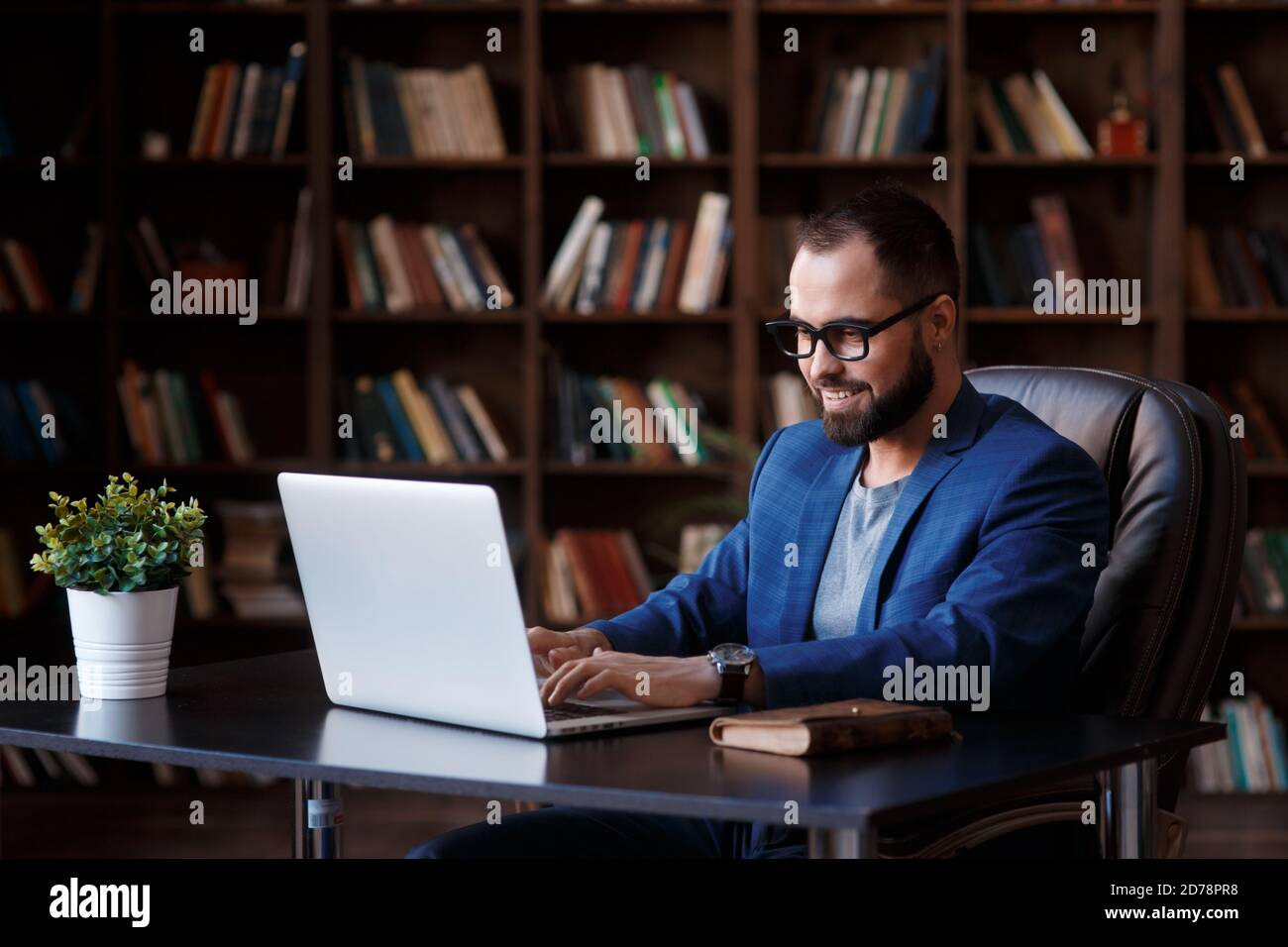 Un giovane uomo d'affari lavora su un computer portatile in un lussuoso ufficio nella biblioteca. Un uomo barbuto e allegro in un vestito blu a una scrivania sta digitando su una tastiera Foto Stock